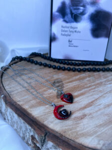 گردنبند ست ماه و قلب با تم هالووین رنگ قرمز و مشکی با زنجیر استیل روی زمینه چوبی کنار کتاب و تسبیح مشکی